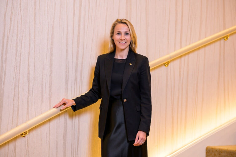 Professional woman in a black blazer and dress standing at a beige striped wall beside a gold handrail on a staircase.