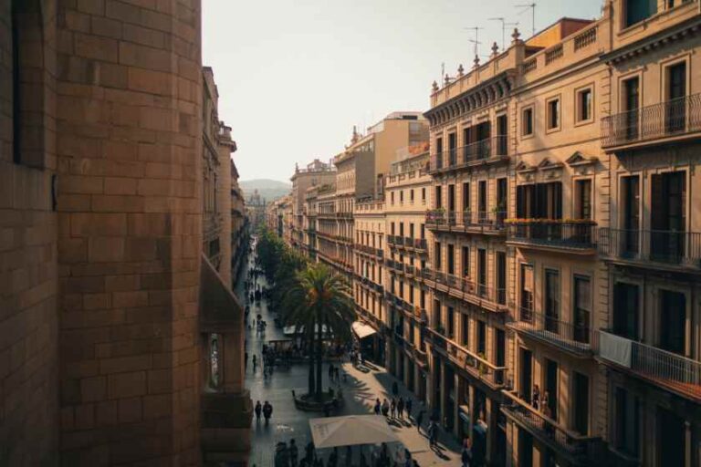 Busy sunlit street in a European city, flanked by tall beige buildings with balconies and a palm tree in the center, pedestrians walking.