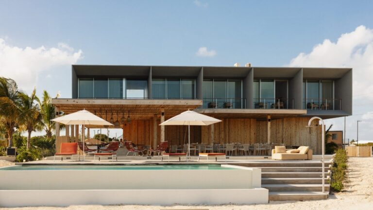 Modern beachfront resort with a wooden poolside deck, white umbrellas, red lounge chairs, and a two-story glass-walled building.