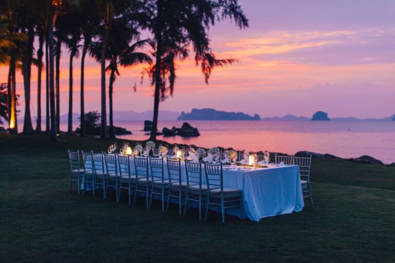Long white-draped banquet table set for a beachside dinner at sunset, with candles and glassware, palm trees in the background.
