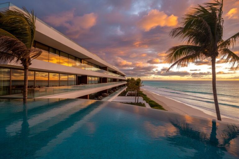 Infinity pool overlooking a sandy beach at sunset, with a modern white resort and palm trees reflecting on the water.