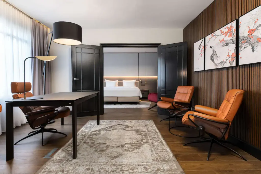 Hotel suite living area with a dark wooden desk, brown leather chair, and warm lighting leading to a bedroom with a bed and side table.