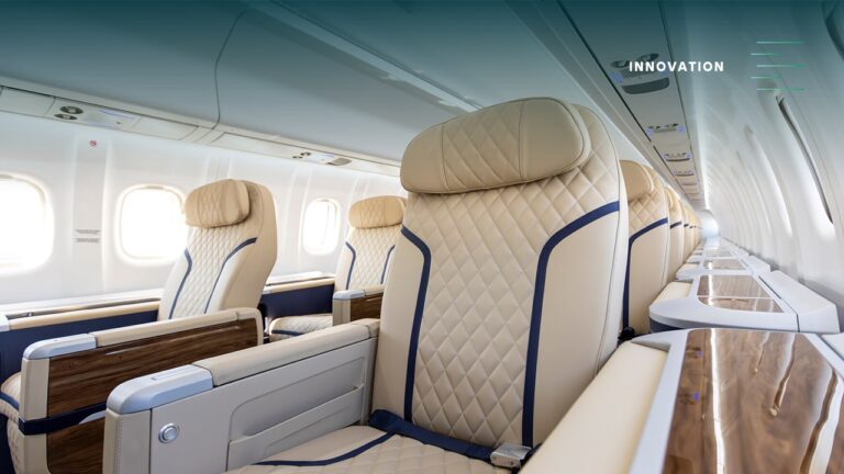 Luxurious aircraft cabin with tan quilted seats, blue piping, and wood-trimmed tables; 'INNOVATION' visible on the ceiling area of the cabin.