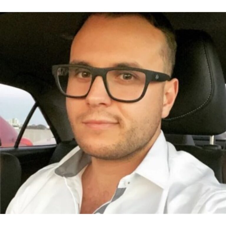 Portrait of a man wearing black-framed glasses, seated in a car and looking at the camera.