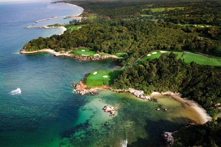 Aerial view of a coastal golf course with green fairways, sand bunkers, and rocky shoreline along turquoise water.