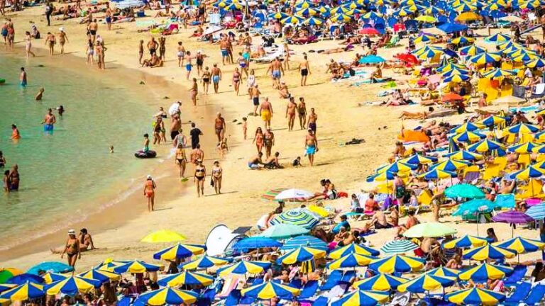 Busy sandy beach crowded with people and blue-yellow umbrellas along the shoreline and in the background