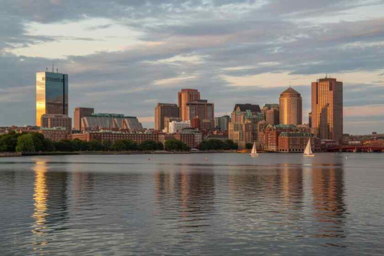 Sunset over a city skyline along a calm river, with several tall buildings reflecting in the water and a few sailboats on the surface.