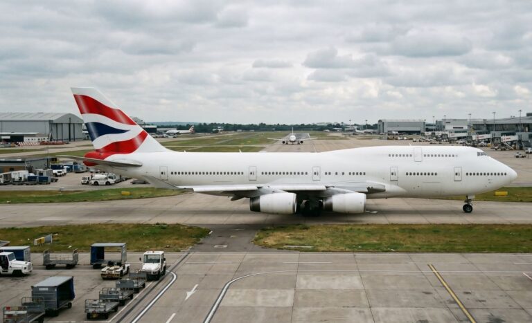 Large white British Airways airliner on a taxiway, red-white-blue tail, airport buildings in the background.