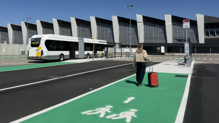 Woman with a red suitcase walking on a green pedestrian lane toward a white bus at a modern bus terminal under a blue sky, daytime.