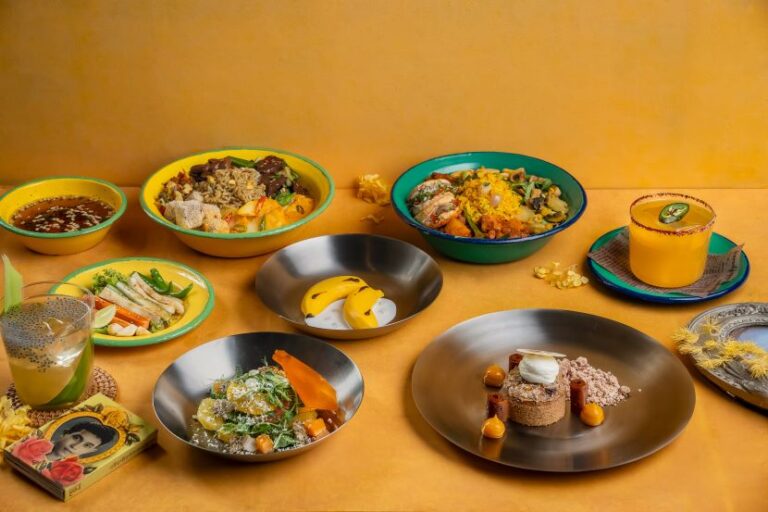 Colorful assortment of bowls with various plated dishes and drinks on a warm yellow table.