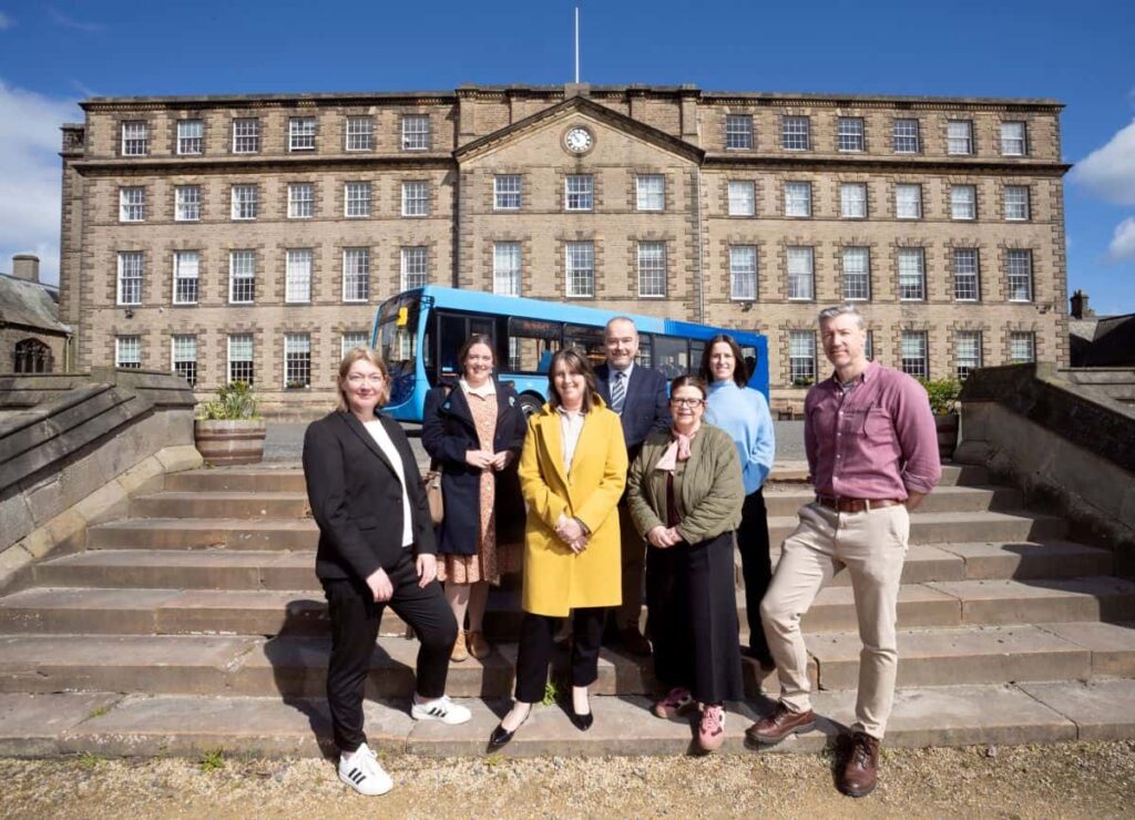 Eight people stand together on stone steps in front of a large brick building with a blue bus behind them.