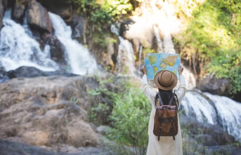Traveler with a brown backpack and hat holds up a map in front of a waterfall by rocks and greenery.