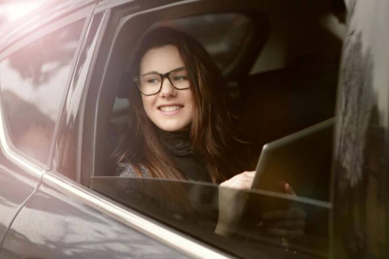 Smiling woman with glasses sitting in a car window, holding a tablet nearby.