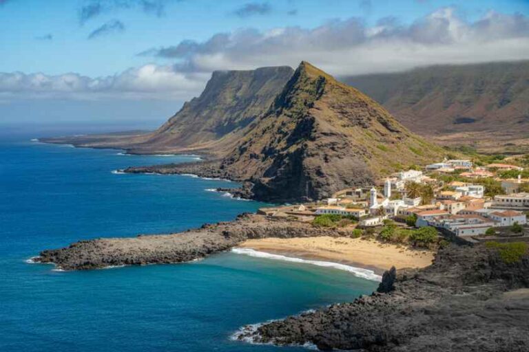Coastal village with white houses at the base of a rugged cliff, overlooking a blue ocean and rocky coastline.