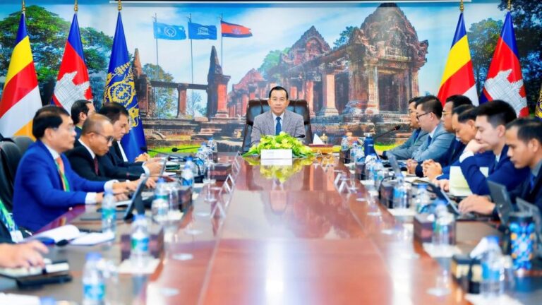 Formal meeting of officials around a long conference table with flags in background and a central speaker at head of table