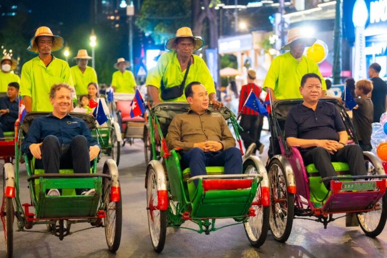 Three men ride in colorful rickshaw-style wheelchairs in a nighttime parade, pulled by attendants in neon yellow shirts; small flags line the chairs as spectators watch.
