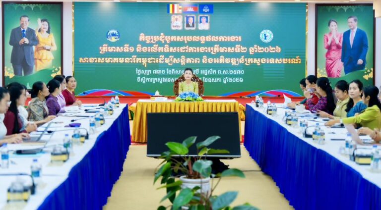 Formal meeting in a conference room with delegates seated along blue-skirted tables around a U shape, a speaker at the head table, and a large green banner behind.