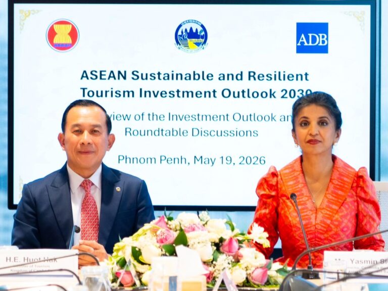 Two panelists, a man in a navy suit and a woman in a red dress, sit at a table with a flower centerpiece during an ASEAN Tourism Investment Outlook event in Phnom Penh, May 19, 2026. Behind them is a large screen with the event title and logos.