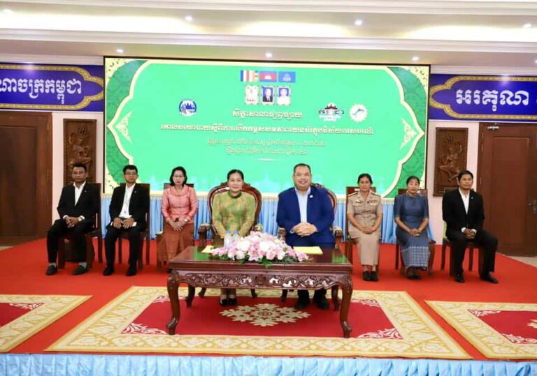 Eight officials sit on a stage with a green backdrops and a central floral centerpiece on a carved table; formal ceremony setting with red carpet and blue skirting.