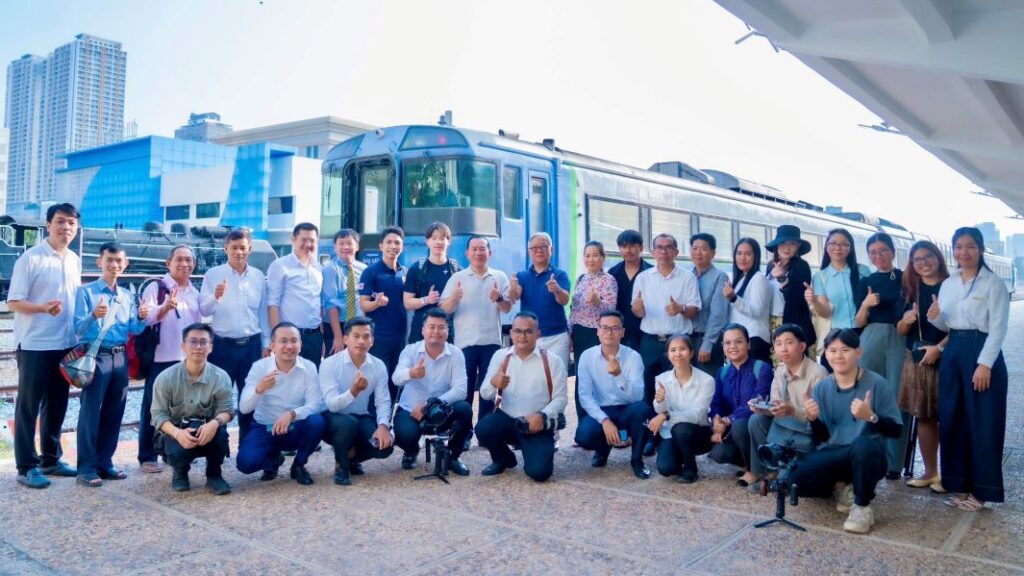 Group of people standing and kneeling, giving thumbs up in front of a blue train at a station (team photo).