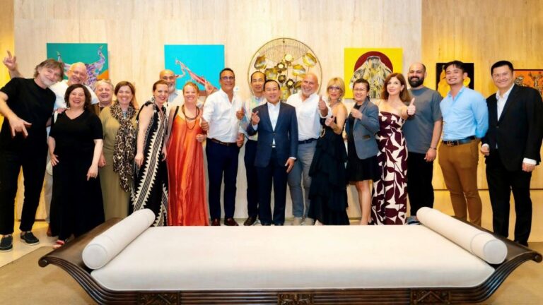 Diverse group of people posing and giving thumbs up in an art gallery, with colorful paintings on the wall behind them and a large decorative bench in the foreground.