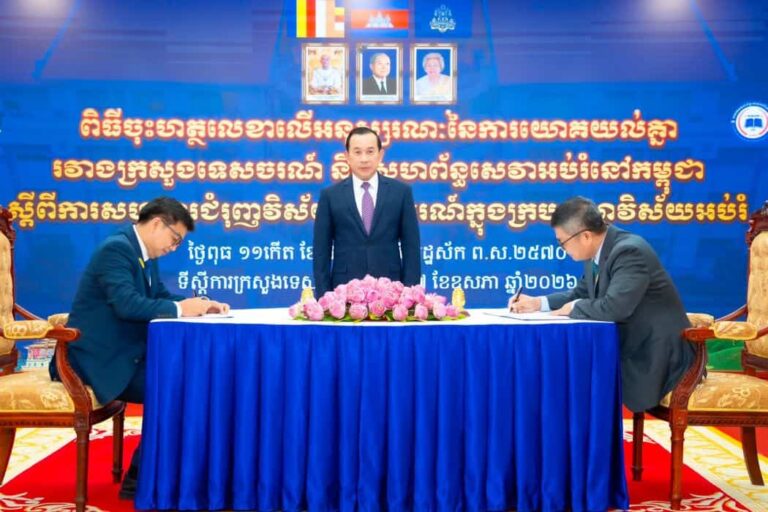 Official signing ceremony: two men sign documents at a blue-draped table, a man stands behind them, with pink flowers and a Khmer backdrop behind.