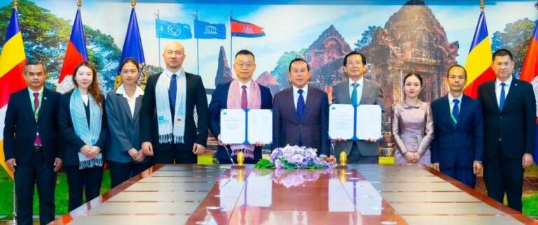 Ten professionals in formal attire stand behind a conference table as two central figures hold signed documents during a formal ceremony, with flags and a scenic backdrop behind them.