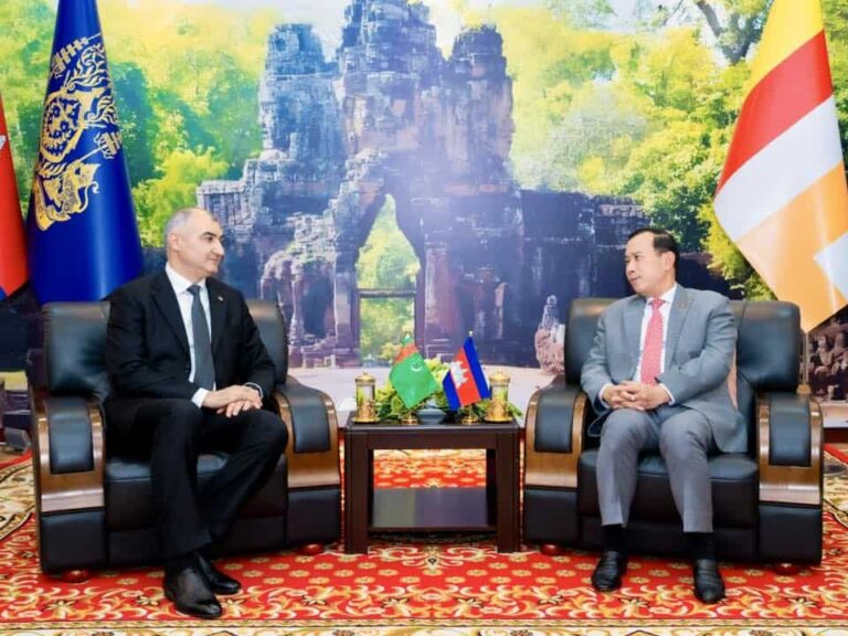 Two men in suits sit in leather chairs across from each other in a formal diplomatic meeting, with flags between them on a small table.