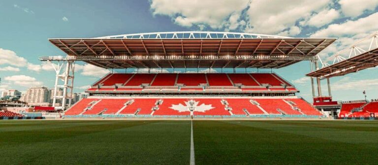 Wide view of a large outdoor stadium with red seating forming a white maple leaf logo and a green pitch under a blue sky with scattered clouds.