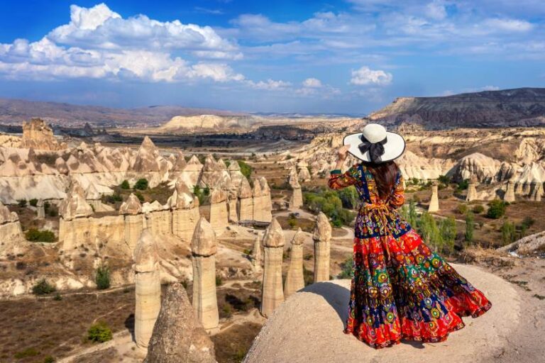 Woman in a bright patterned dress and wide hat stands on a rocky overlook, viewing Cappadocia's fairy chimneys and desert landscape.