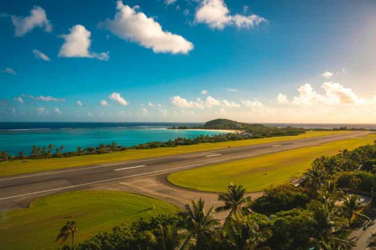 Aerial view of a tropical coastline with a runway bordered by green grass and palm trees, and turquoisesea beyond the coast.