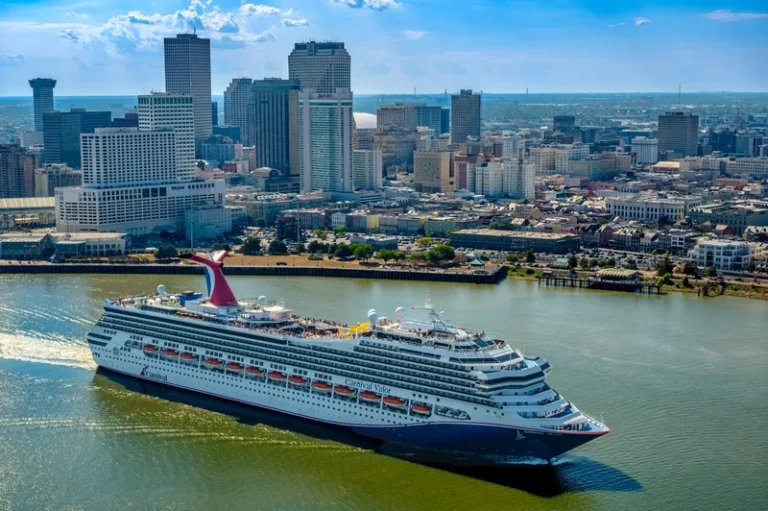 Carnival Valor cruise ship sailing past a city skyline along a river on a sunny day.