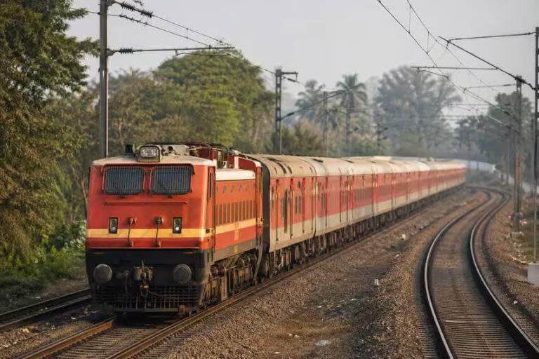 Red passenger locomotive pulls a long red-and-white train on a curved track through a green, tree-lined area with overhead electric lines nearby.