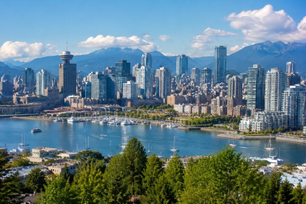 City skyline along a blue harbor with boats, framed by green trees in the foreground and mountains in the distance.