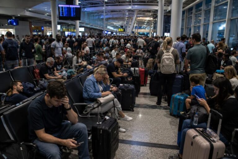 Crowded airport terminal with travelers seated and standing, many with luggage, waiting near gates and screens showing flight information.