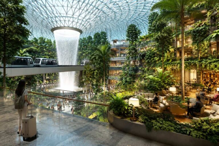 Lush indoor atrium with a towering waterfall under a glass geodesic dome, a suspended train on a bridge, and people seated in a garden lounge area.