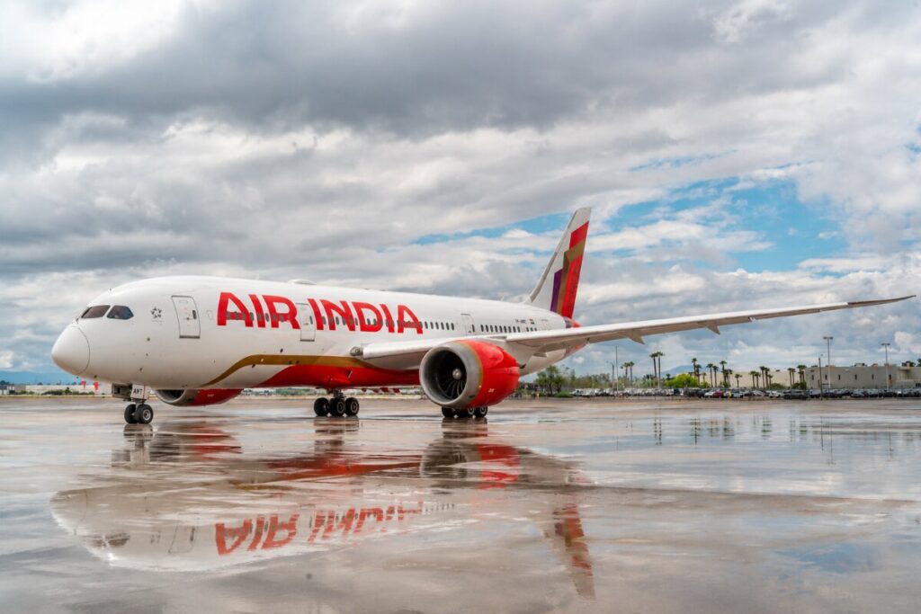 Air India jet parked on a wet tarmac with reflections, under a cloudy sky and distant airport buildings in the background.
