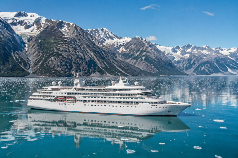 White luxury cruise ship on calm glacier-blue water with snow‑capped mountains in the background.