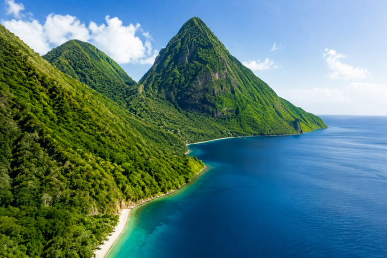 Lush green mountains meet a turquoise bay with a narrow sandy beach along the coast under a blue sky.