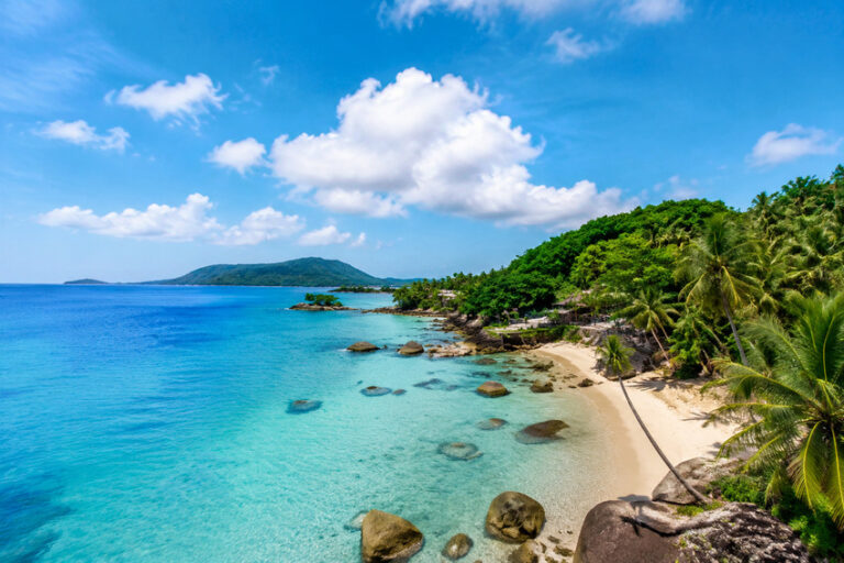 Tropical beach with clear turquoise water, sandy shore, and palm trees along a rocky coastline under a blue sky with clouds.