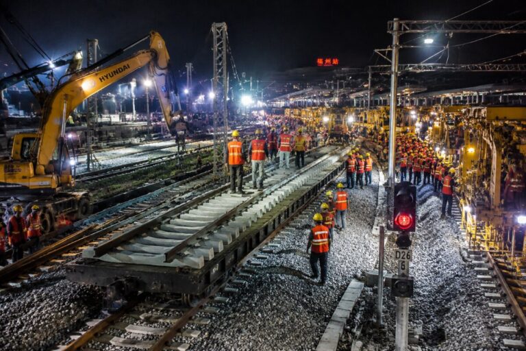 Nighttime railway construction site with workers in orange vests guiding machinery laying concrete sleepers on tracks under bright lights.