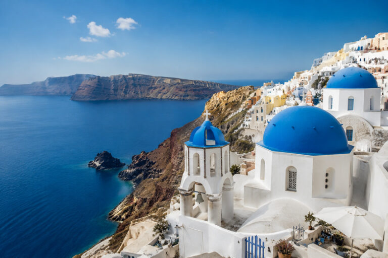 White-walled buildings with blue domes cascade along a cliffside overlooking the deep blue Aegean Sea in Santorini, Greece