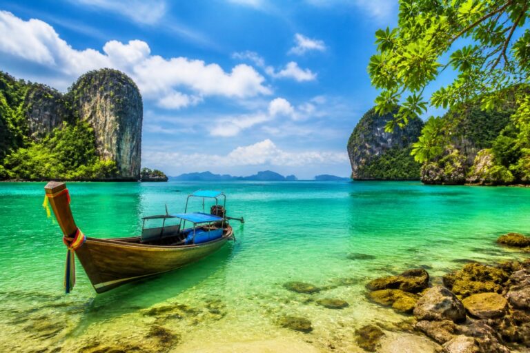 Traditional wooden boat anchored on a clear turquoise beach, with lush cliffs and blue sky overhead.