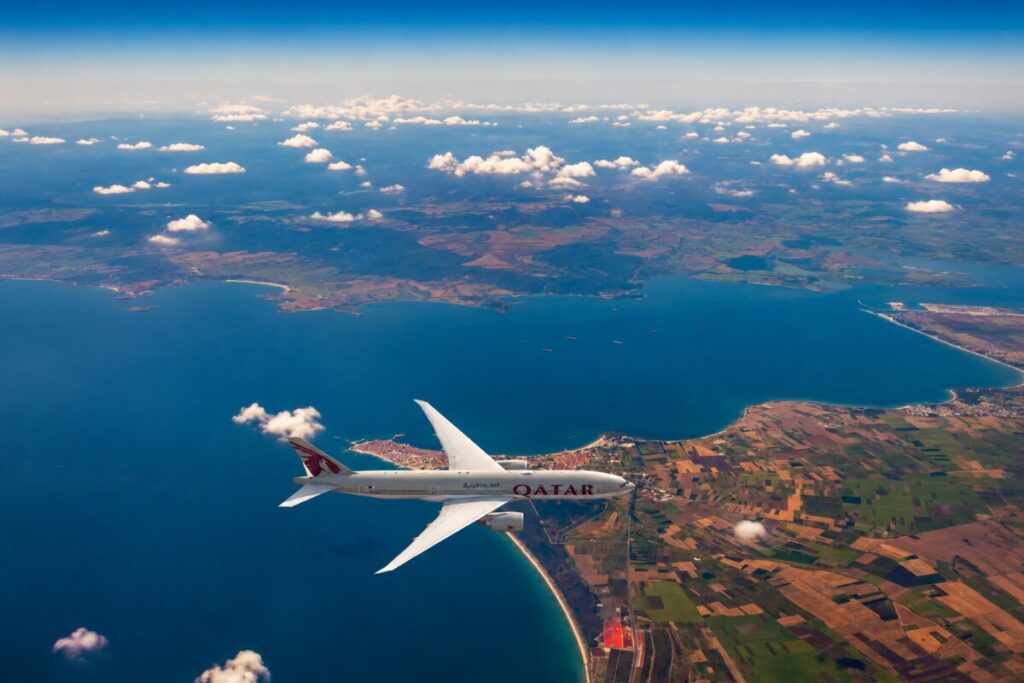 Qatar Airways airplane cruising above a blue sea and patchwork coastline during daytime over clouds.