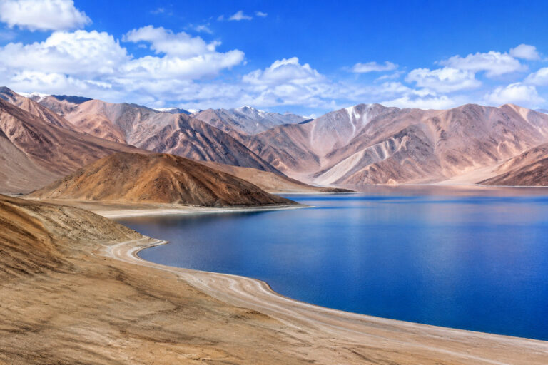 A deep blue lake curves along a sandy shoreline with brown, barren mountains and a bright blue sky above.