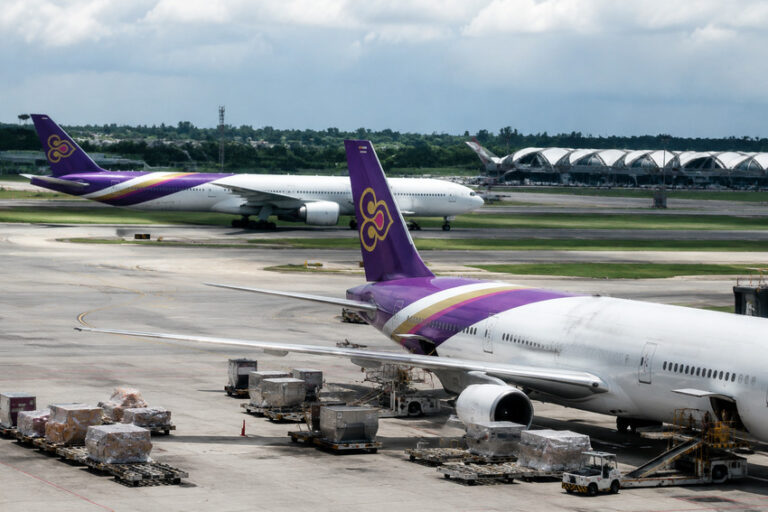 Two commercial airliners on an airport apron; foreground plane has purple and gold livery with cargo pallets nearby, a second plane in the background.