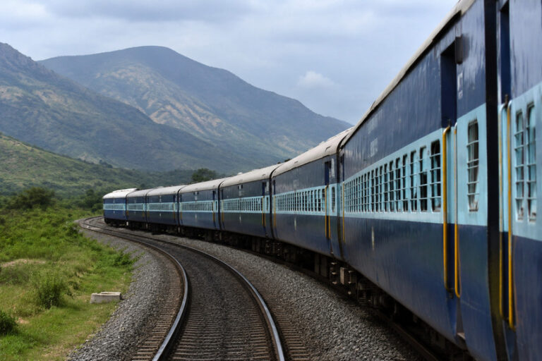 Blue passenger train curving along tracks through green countryside with mountains in the distance under a cloudy sky.