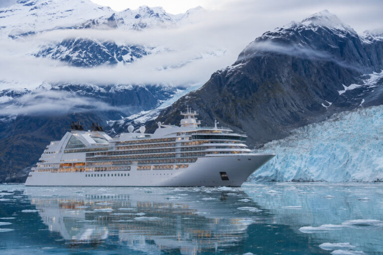 Large white cruise ship sailing near a blue glacier in a fjord with snow-covered mountains behind it.