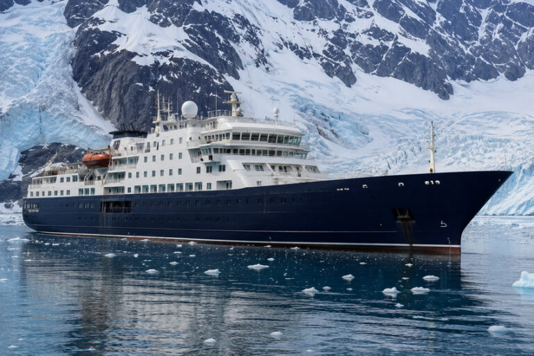 Large dark-blue cruise ship sailing through icy Arctic waters with a glacier and snow-covered mountains in the background.