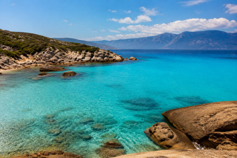 Turquoise sea along a rocky coastline with clear water and distant mountains under a blue sky.